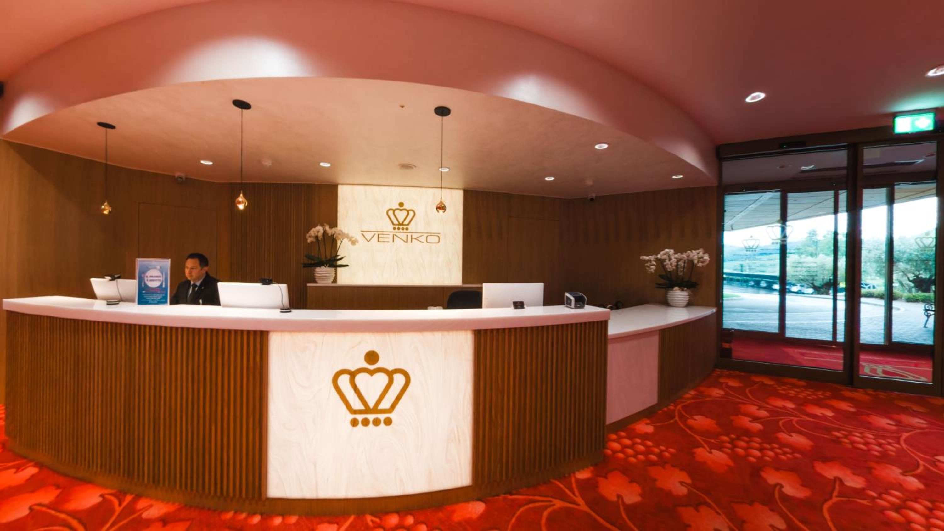 A reception desk with a logo, a man seated behind it, and a view of the outdoors through large windows.