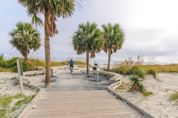A wooden boardwalk leading to a sandy beach area with palm trees and benches.