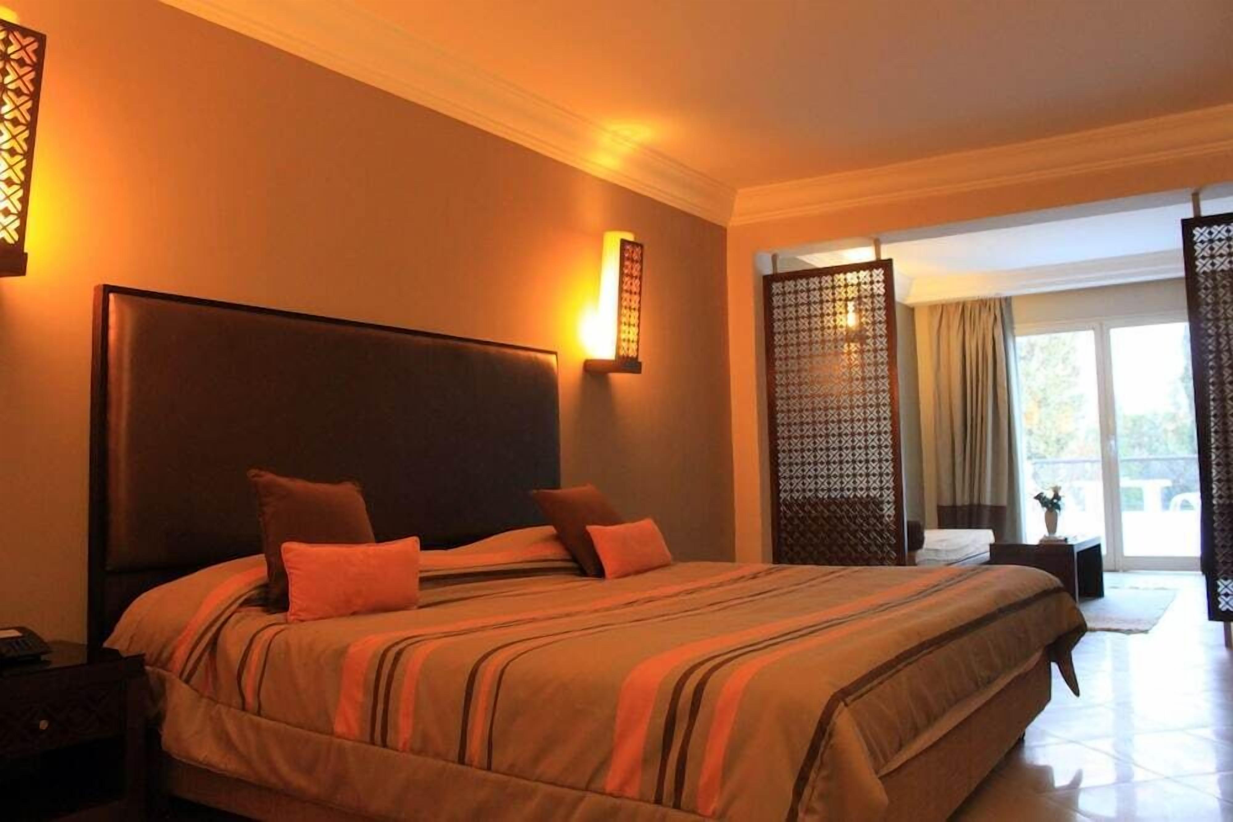 A hotel room with a large bed, a nightstand, a wall-mounted lamp, and a view of the outdoors through a sliding door.