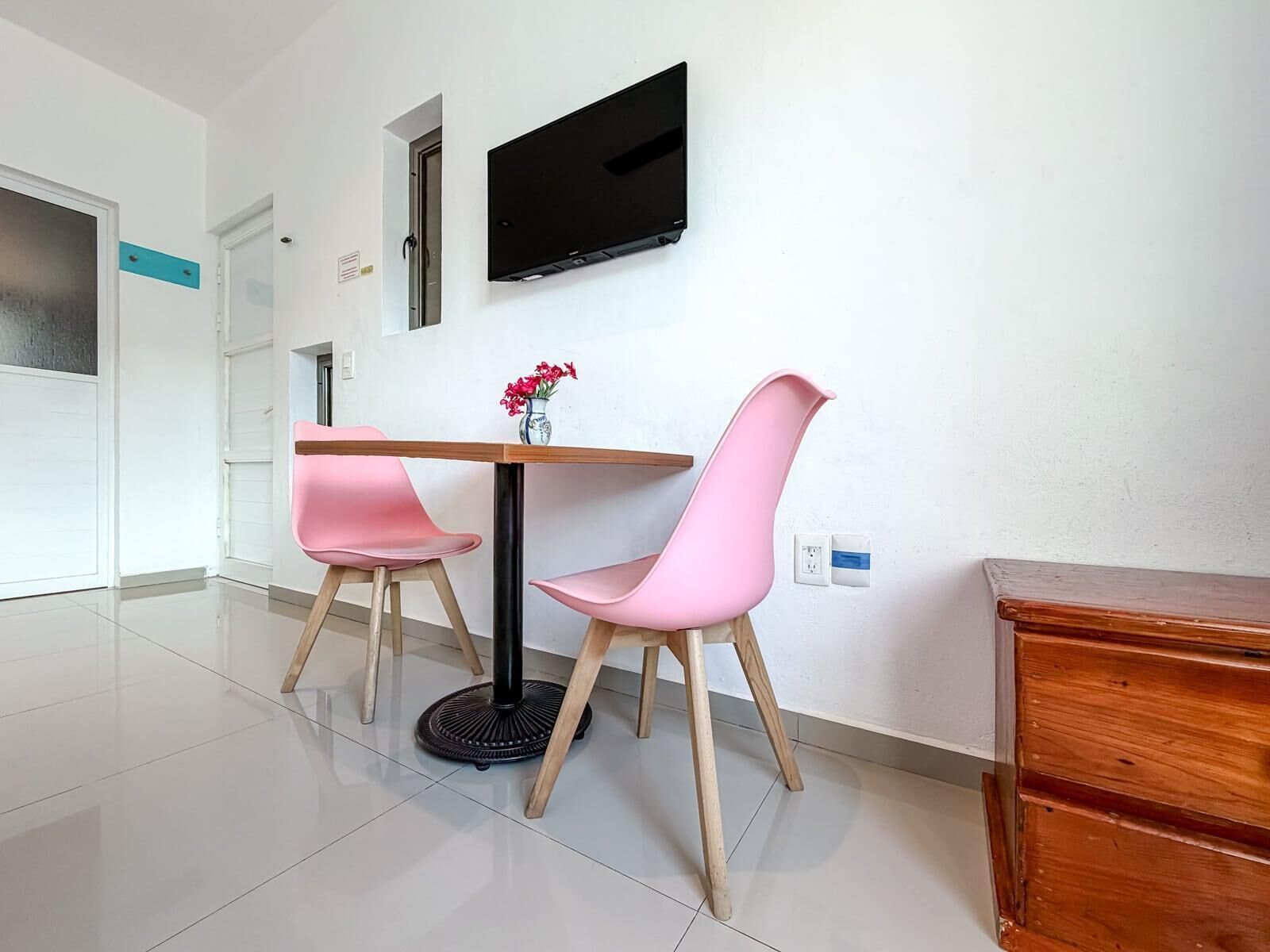 Une pièce moderne avec une table en bois, deux chaises roses, un pied de table rond noir, un téléviseur à écran plat fixé au mur et un meuble en bois.