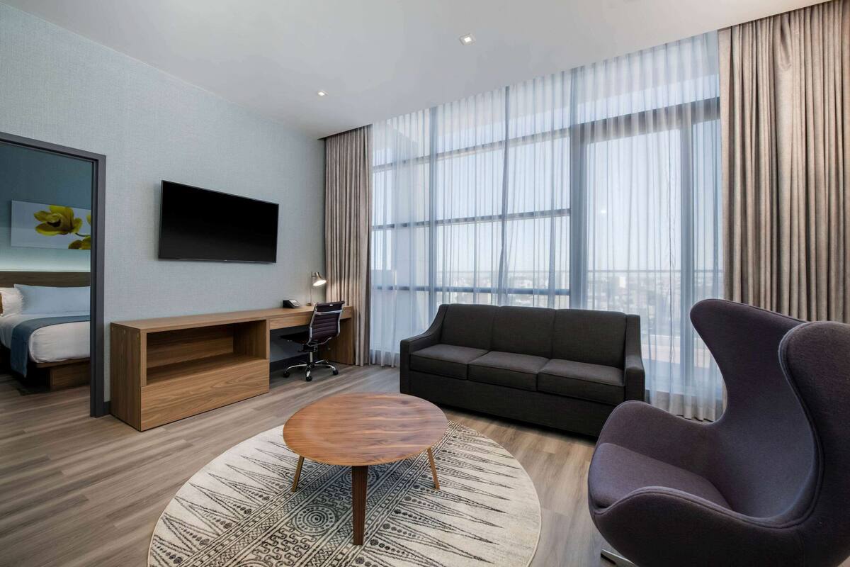 Une chambre d’hôtel moderne dotée d’une grande fenêtre, d’un téléviseur à écran plat, d’un canapé, d’un fauteuil, d’un bureau et d’un lit.