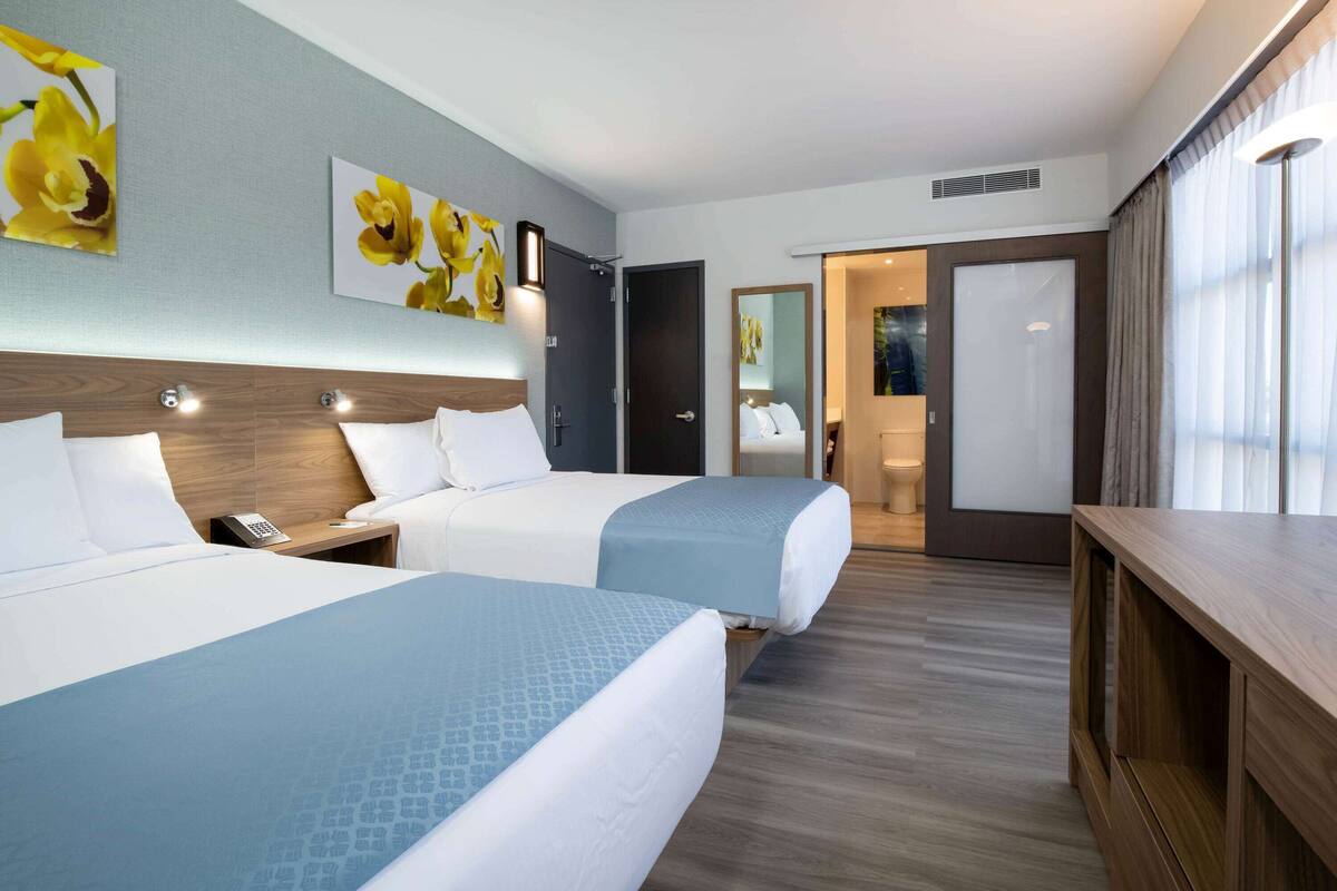 Une chambre d’hôtel avec deux lits, une tête de lit en bois, un bureau et une grande fenêtre avec des rideaux.