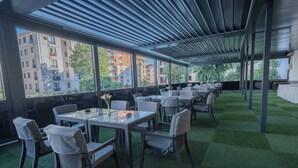 A covered outdoor dining area with tables and chairs, large windows, and a view of buildings. - Tryp By Wyndham Istanbul Sancaktepe (Istanbul)