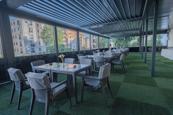 A covered outdoor dining area with tables and chairs, large windows, and a view of buildings. - Tryp By Wyndham Istanbul Sancaktepe (Istanbul)