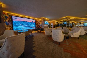 Un salon d’hôtel doté de sièges confortables, d’un grand écran de télévision diffusant des images d’une ville, et d’un mur décoratif orné de noms de villes.