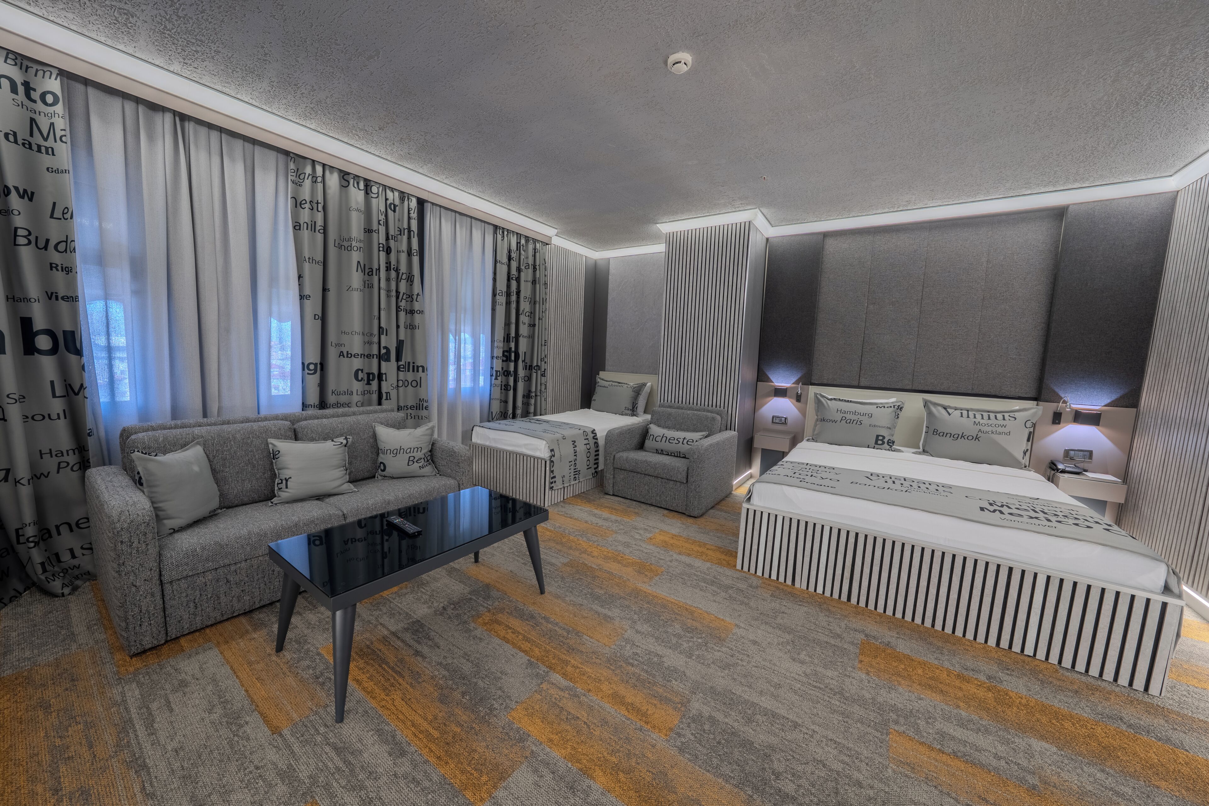 A modern hotel room with a sofa, a small table, a bed with a headboard, and a window with curtains.