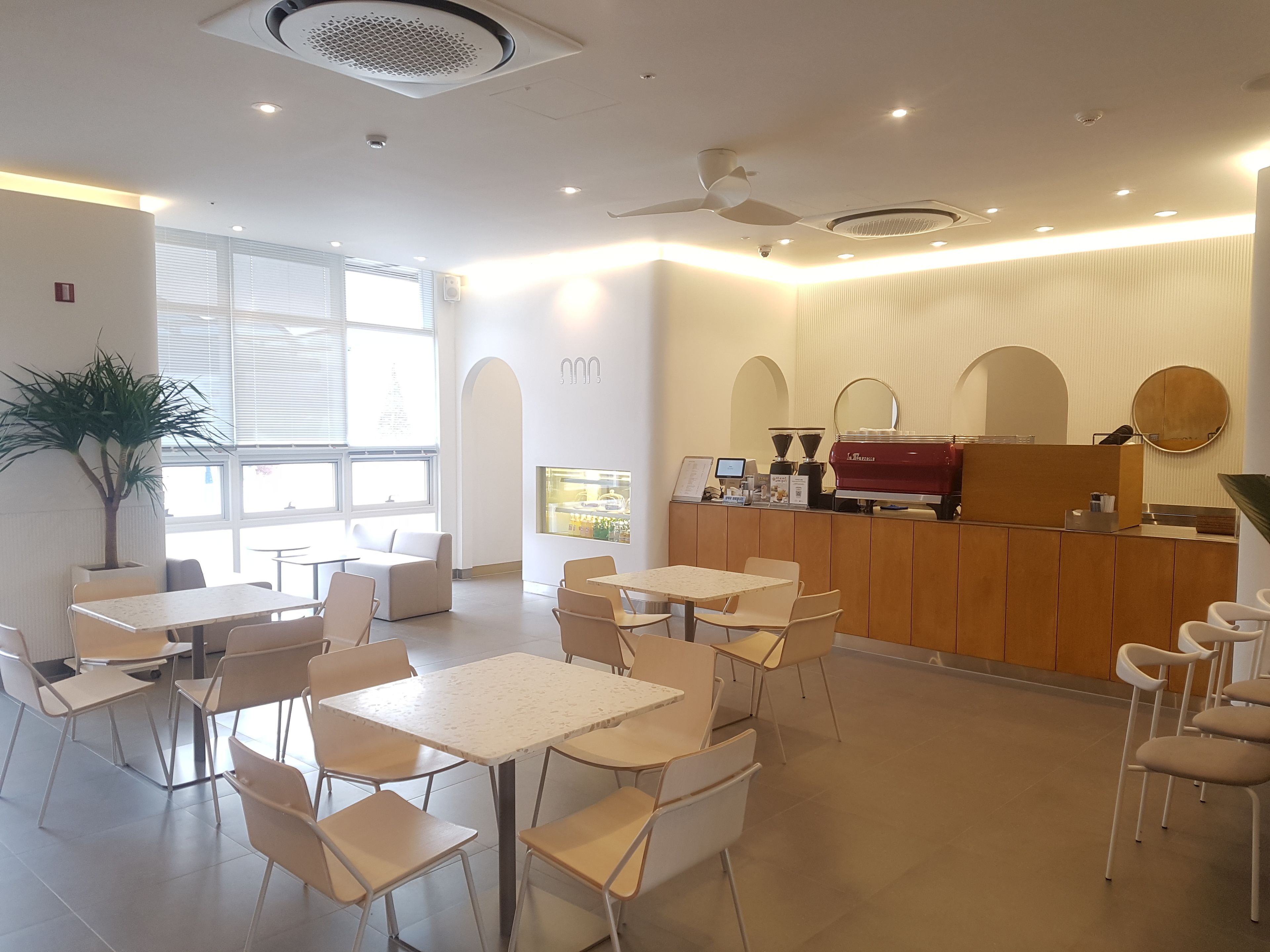 Una cafetería moderna con mostrador de madera, varias mesas y sillas, y un amplio ventanal que proporciona luz natural.
