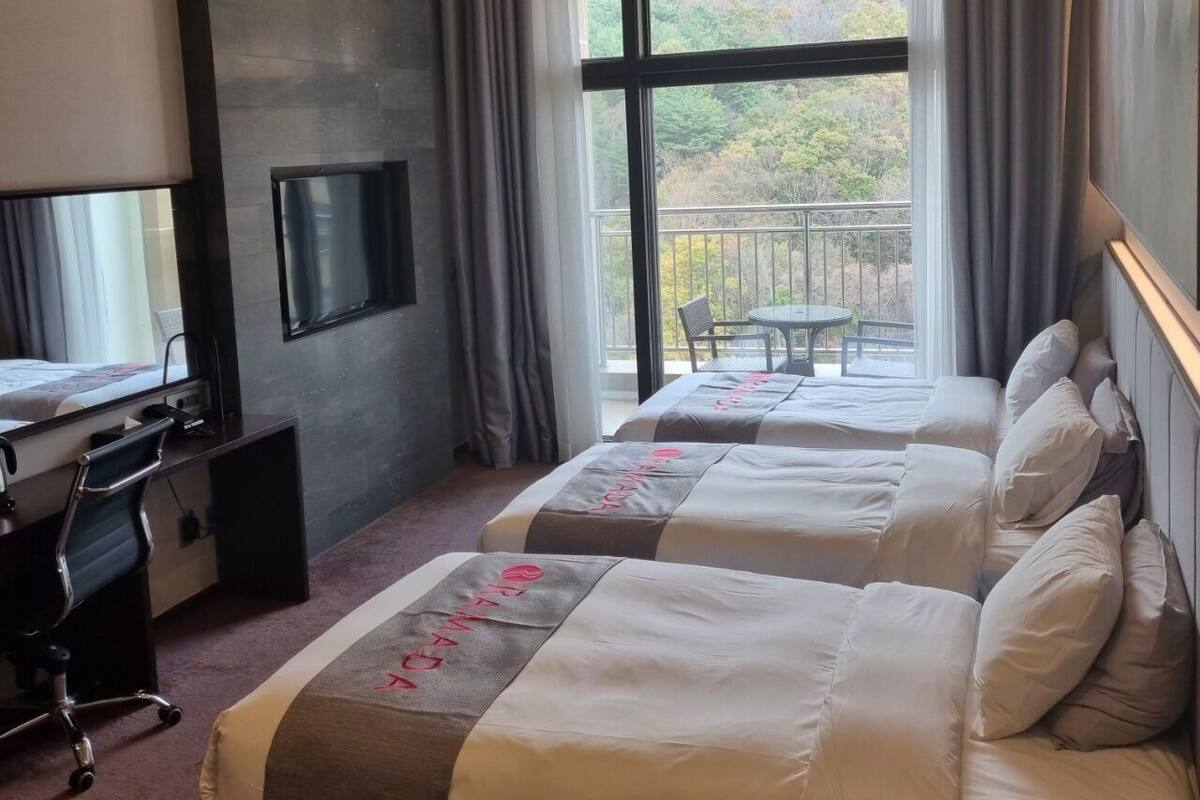 A hotel room with two beds, a desk with a chair, a TV, and a balcony with a table and chairs.