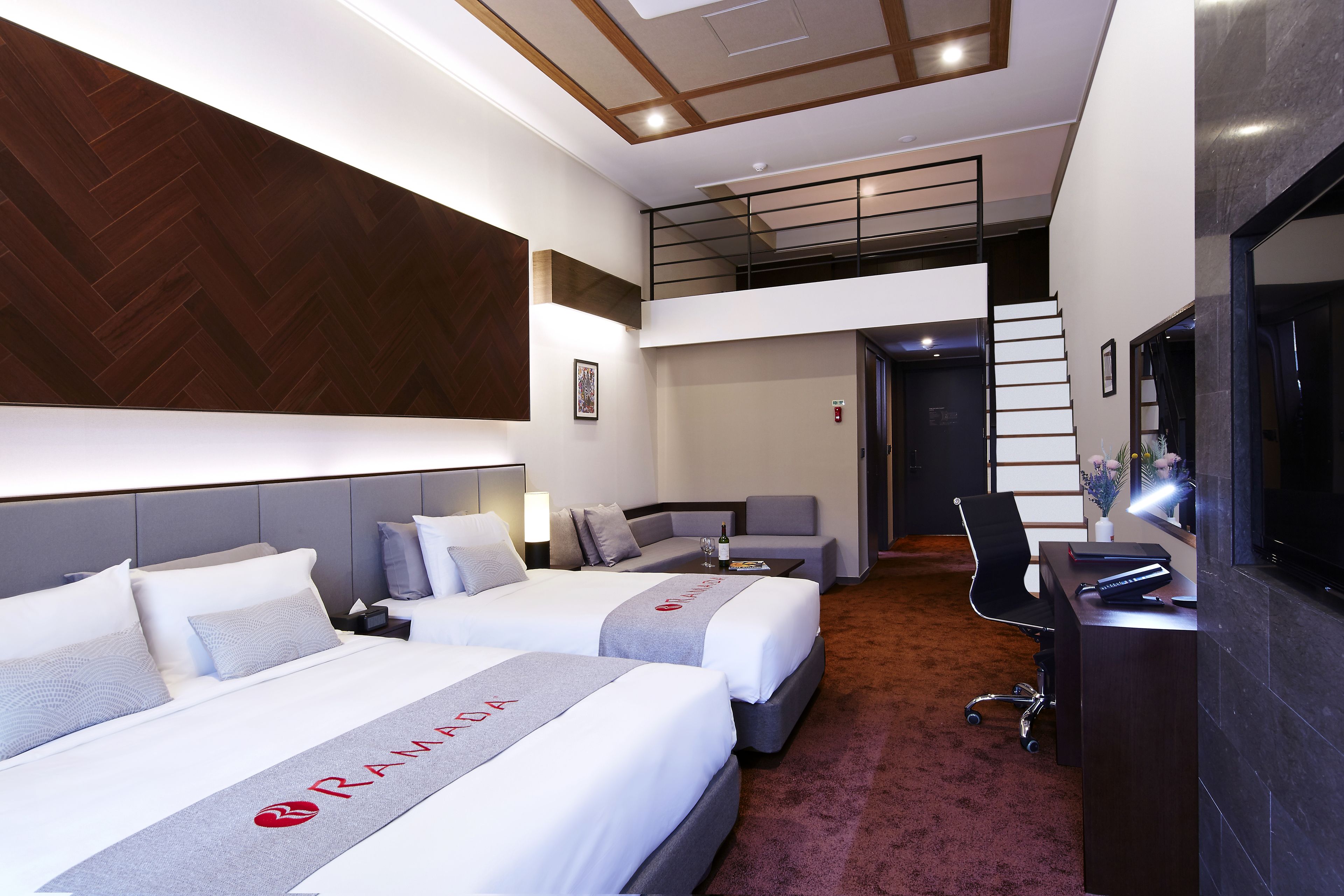 A hotel room with two beds, a desk, a chair, a staircase, and a TV.