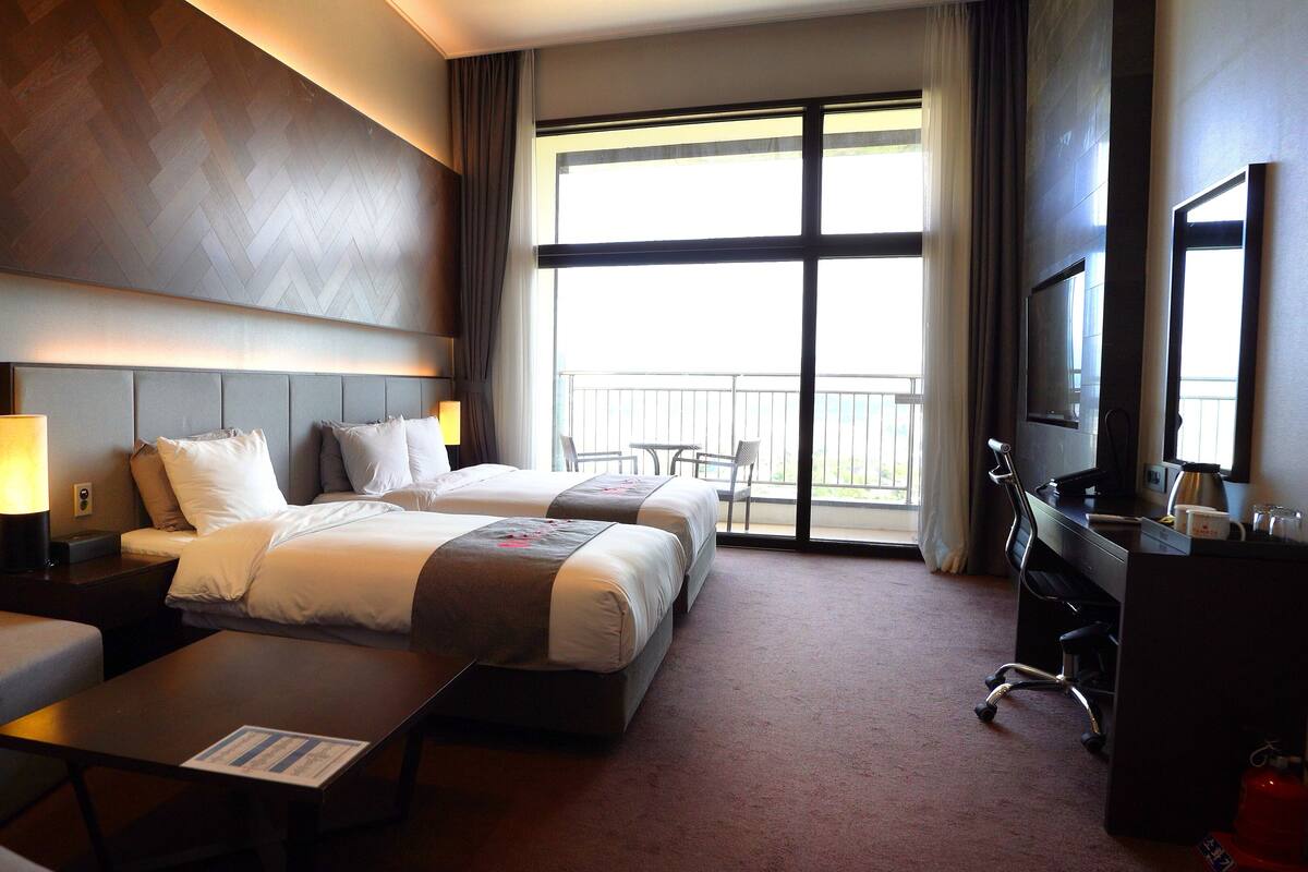 A hotel room with two beds, a sofa, a desk, a TV, and a balcony with a view.