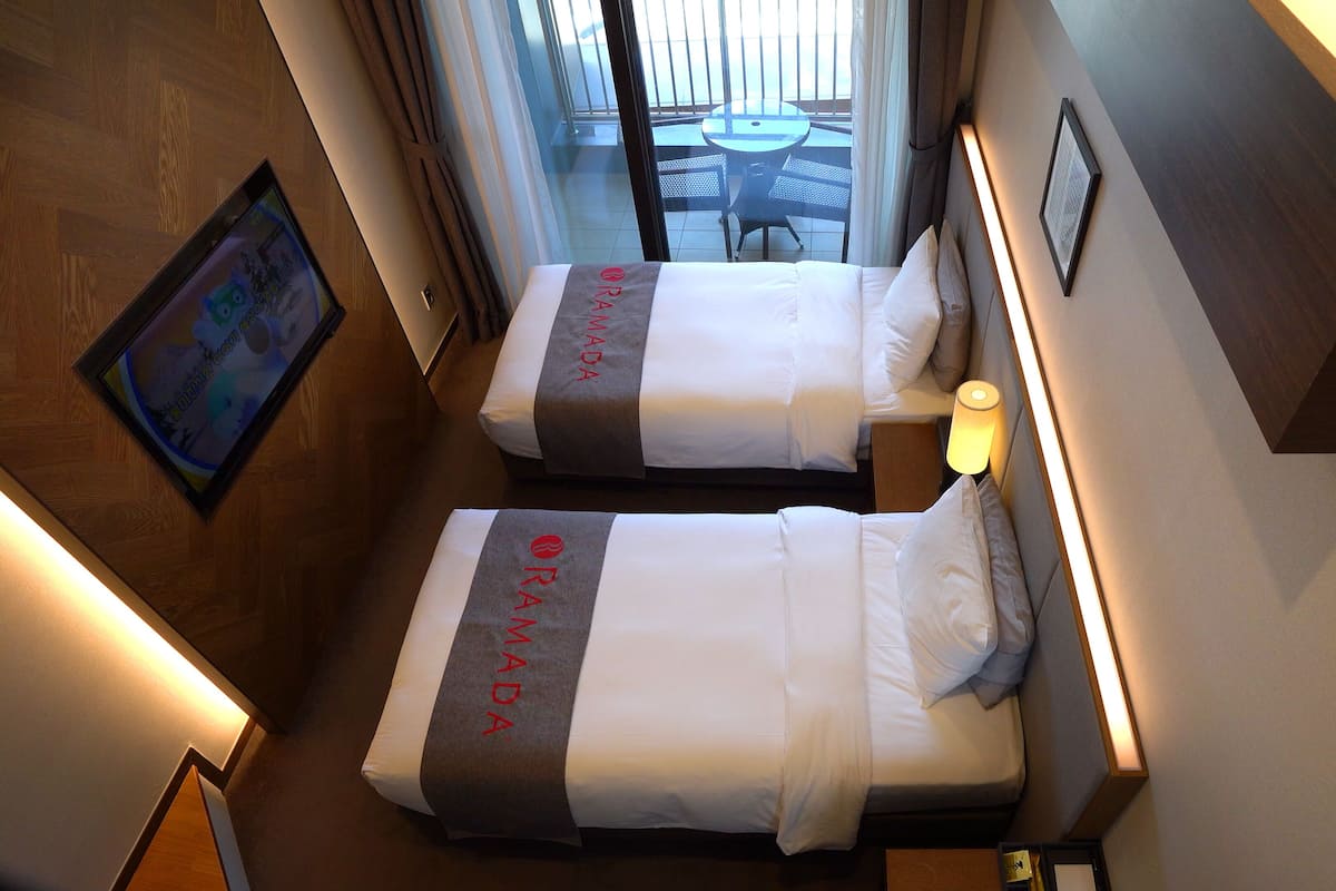 A hotel room with two beds, a small table, a wall-mounted TV, and a balcony with a table and chairs.