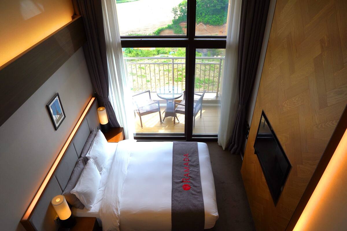 A hotel room with a bed, bedside lamps, a view of the outdoor balcony with chairs and a table, and a framed picture on the wall.