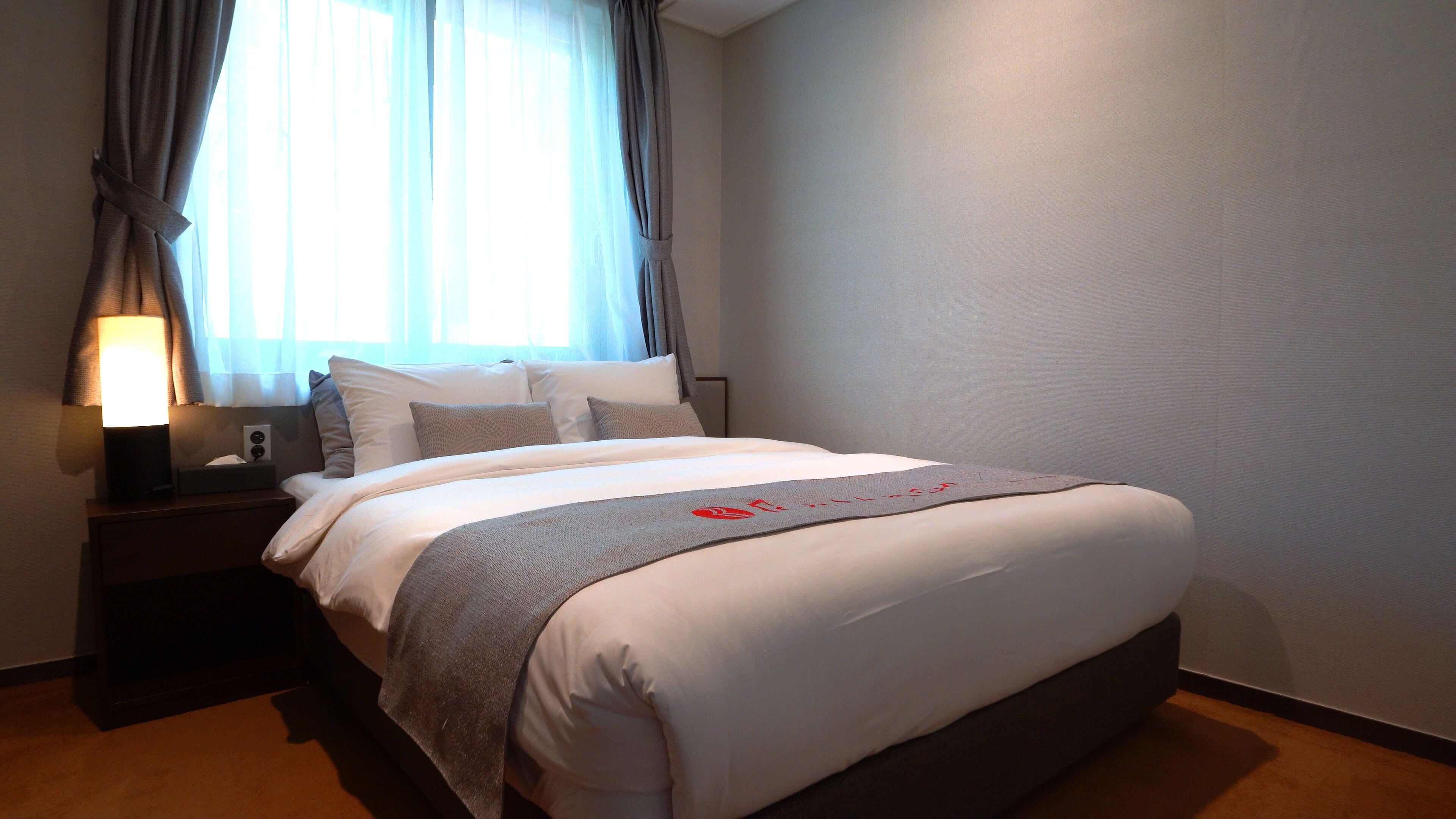 A neatly made bed with a white comforter and a grey throw blanket, a bedside lamp, and a nightstand with a tissue box.