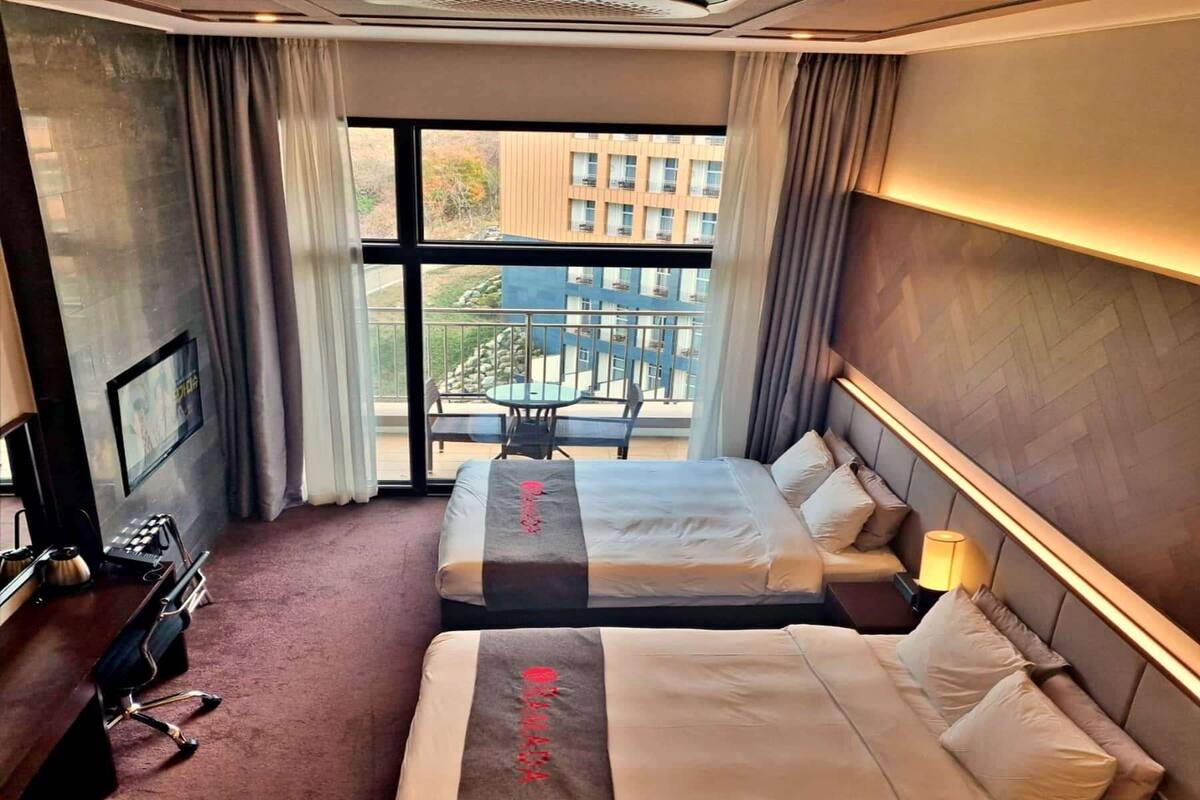 A hotel room with two beds, a desk, a chair, and a balcony with a view of buildings.