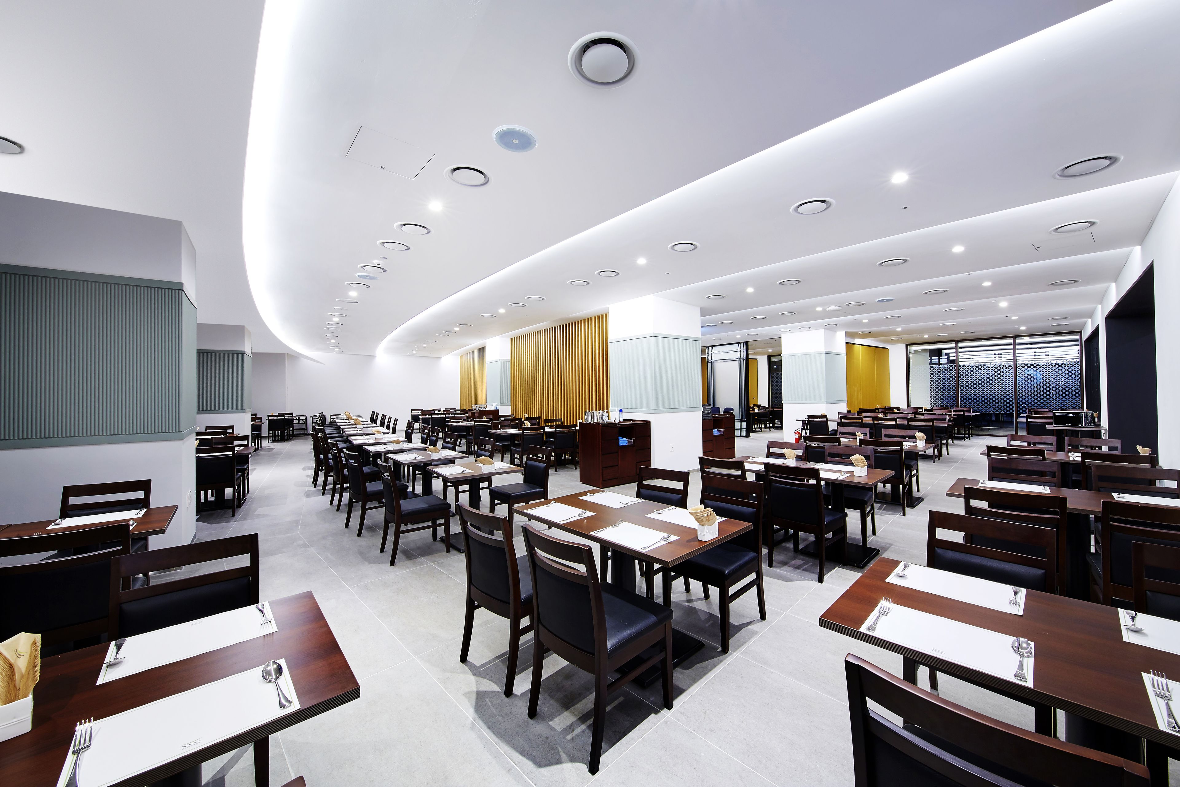 A spacious dining area with multiple tables and chairs, a modern ceiling design, and a large window providing natural light.