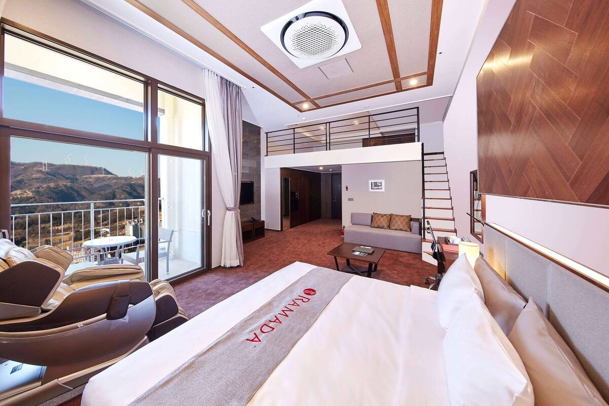 A hotel room with a large bed, a massage chair, a balcony with a view of wind turbines, and a small seating area.