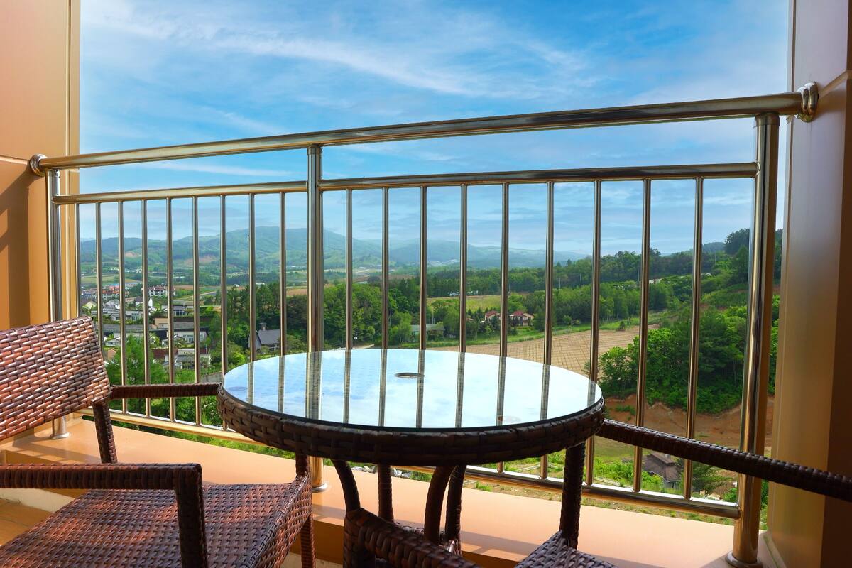 A balcony with wicker furniture and a glass table, overlooking a scenic landscape with houses and greenery.