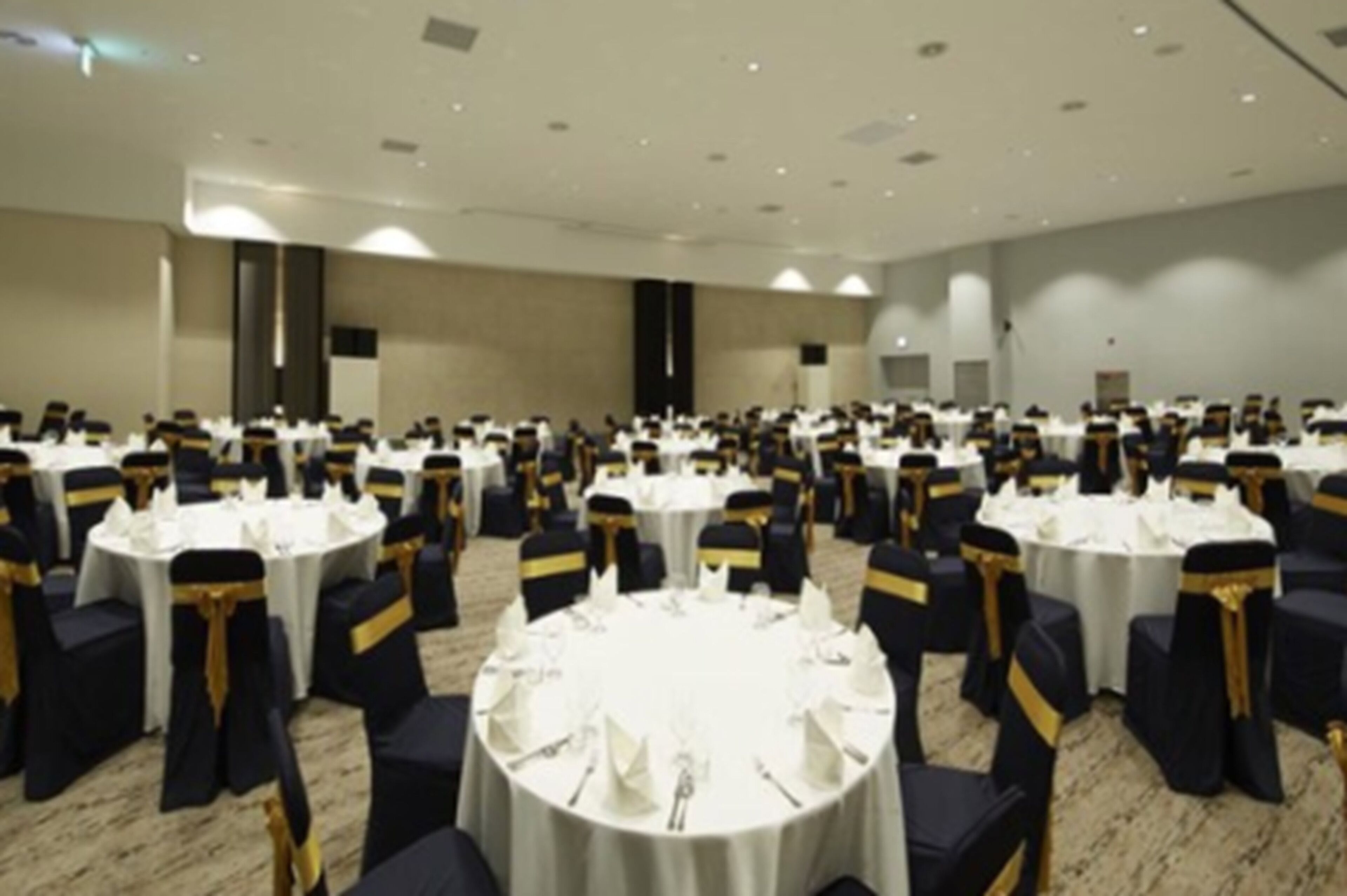 Un amplio salón de banquetes con mesas redondas dispuestas para cenar, cada una con manteles blancos y sillas negras adornadas con cintas doradas.