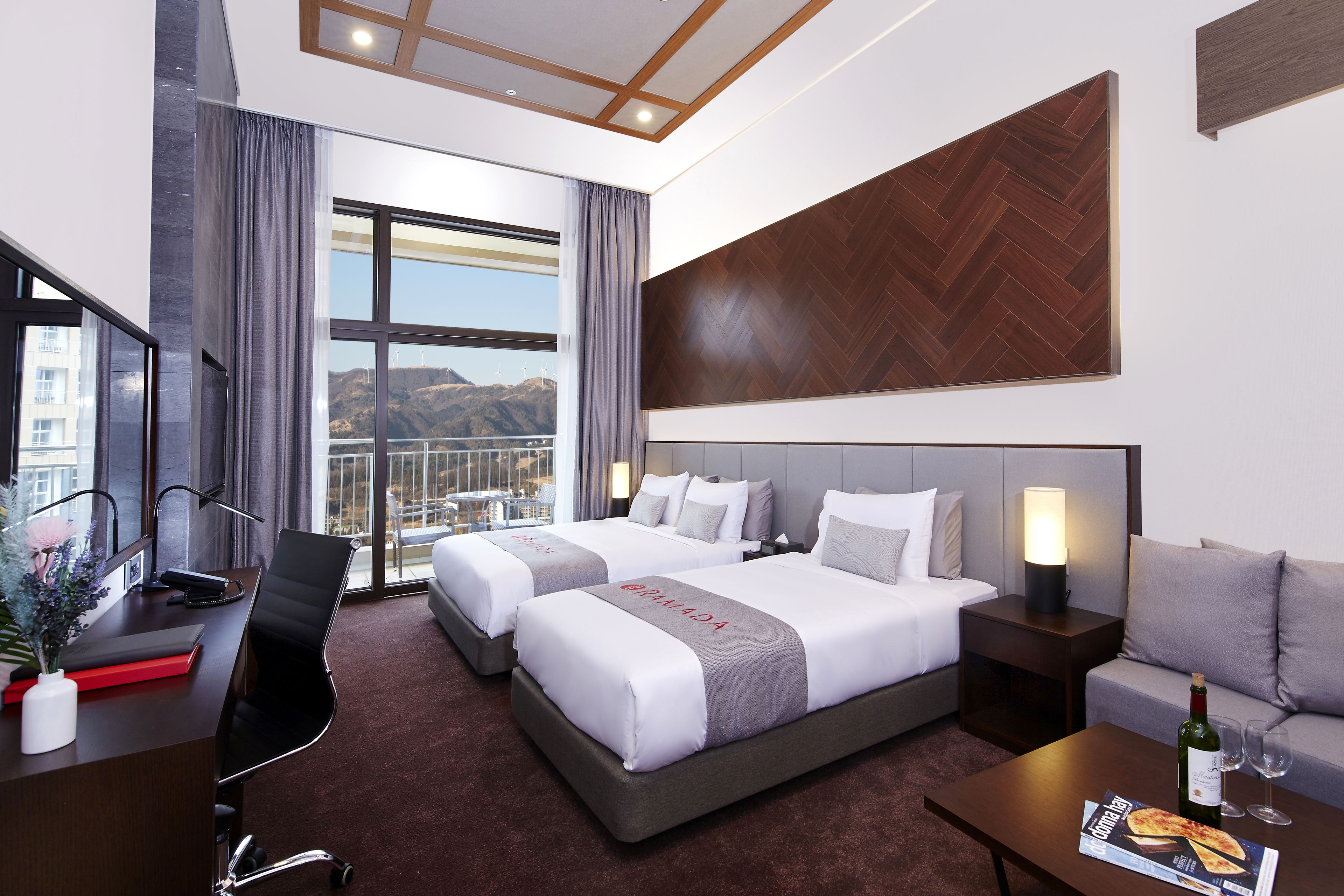 A hotel room with two beds, a desk, a chair, a sofa, a TV, and a window with a view of mountains.