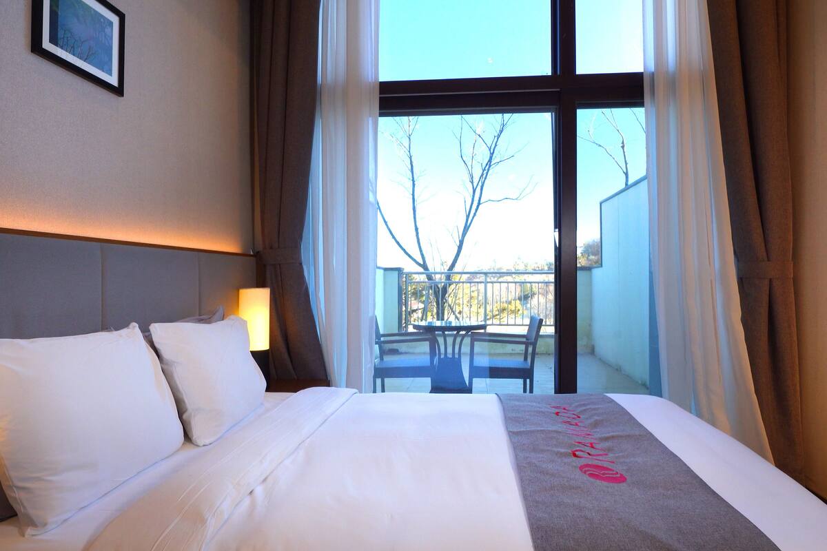 A hotel room with a large bed, a nightstand, a lamp, and a view of the outside balcony.