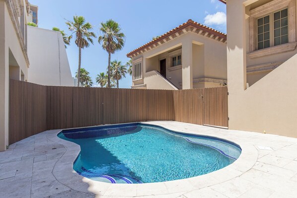 Una piscina con cerca de madera, rodeada por un patio con baldosas y un edificio con techo de tejas.