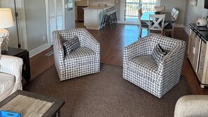 A spacious living room with a large window, a dining area with a table and chairs, and a kitchen in the background. - Canal/w dock, pool, ocean view, 1 block from beach & EV charger (Ocean Isle Beach)