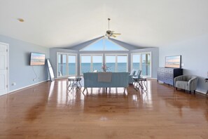 Une pièce spacieuse dotée d’une grande baie vitrée donnant sur la mer, d’un ventilateur de plafond et d’une table à manger avec des chaises.