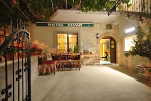 Entrada d'un hotel amb una porta de ferro forjat, una banca amb coixí estampat i una taula amb caires.