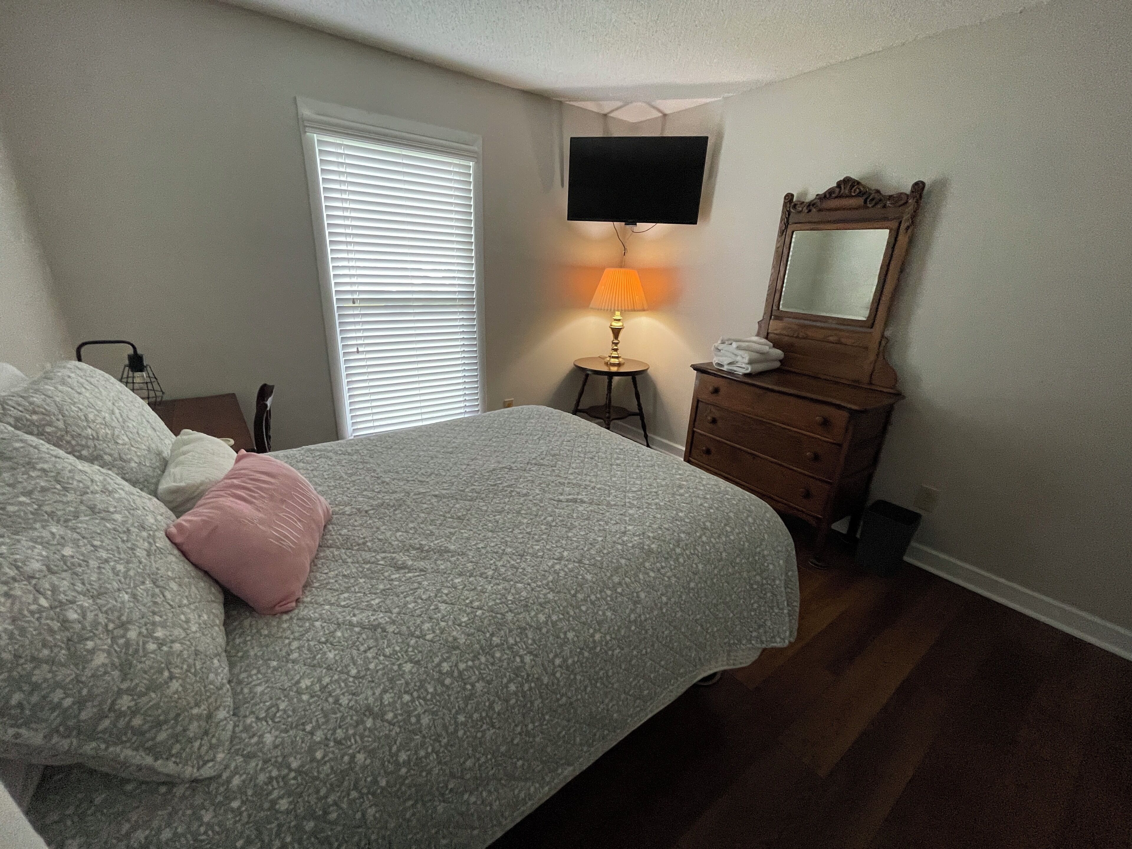 A bedroom with a bed, a dresser, a television mounted on the wall, and a window with blinds.