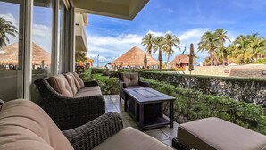 A balcony with wicker furniture, a table, and a view of a resort with palm trees and thatched-roof huts.