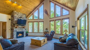 A spacious living room with a stone fireplace, leather sofas, a wooden coffee table, and large windows offering a view of the outdoors.