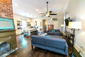 A living room with a fireplace, a sofa, a dining area with a kitchen in the background, and a ceiling fan.