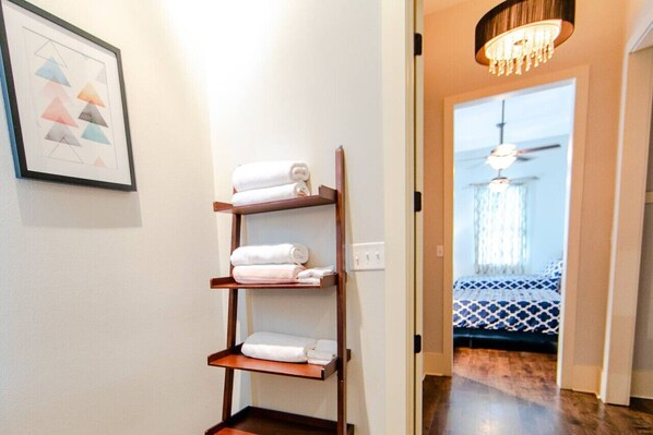 A hallway with a wooden shelf holding towels, a chandelier, and a bed with a patterned bedspread in the background.