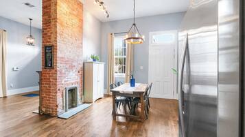 A kitchen with a brick wall, wooden flooring, a dining table with chairs, a refrigerator, and a hanging light fixture.