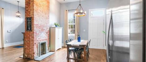 Una cocina con pared de ladrillo, suelo de madera, una mesa de comedor con sillas, un refrigerador y una lámpara colgante.