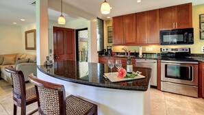 A modern kitchen with wooden cabinets, a granite countertop, a built-in oven, and a microwave. There is a bar area with two chairs and a bottle of wine. - Wailea Elua #207 Ocean and Garden View Short Walk to the BEACH ..Not 14 Min (Wailea)