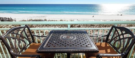 A table and chairs on a balcony overlooking a beach with a fence and a pier in the background.