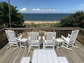Une terrasse en bois avec des chaises Adirondack blanches et une table, donnant sur une plage et l’océan.