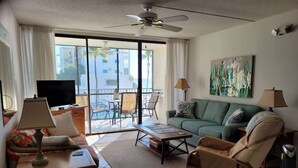 Un salon comprenant un canapé, un fauteuil, une table basse et un téléviseur. Il y a un balcon d’où l’on peut admirer les bâtiments et les palmiers.