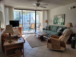 A living room with a sofa, armchair, coffee table, and TV. There is a balcony with a view of buildings and palm trees.