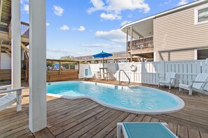 En træterrasse med swimmingpool, liggestole og et overdækket område med en blå parasol.