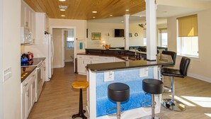 A modern kitchen with a central island, bar stools, and a dining area with a view. - "Sandy Dunes" - Oceanfront Home w/ Private Pool, 8 Bedrooms / 6.5 Bathrooms (Holden Beach)