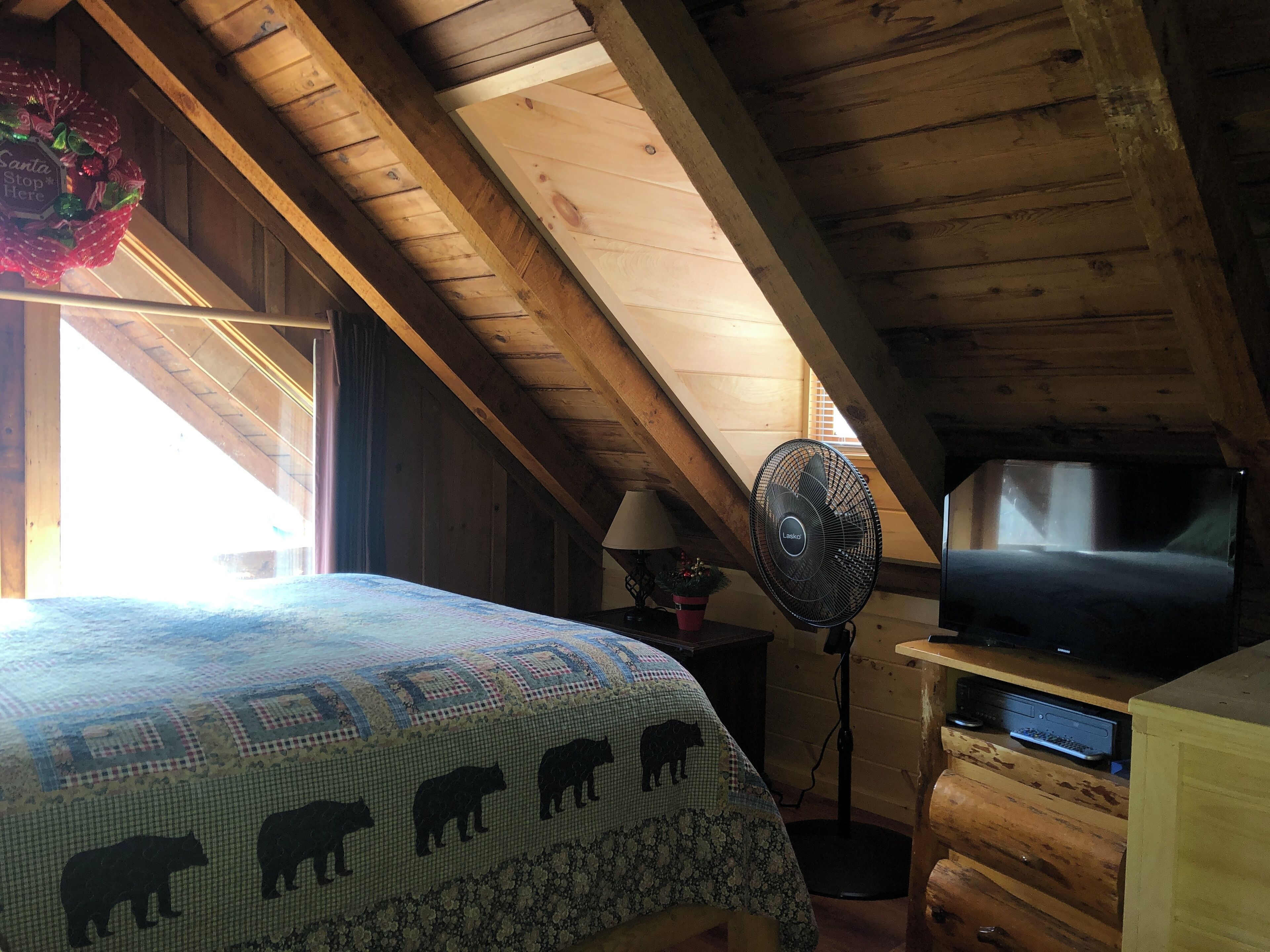 Una camera da letto mansardata accogliente, con un letto, una ventola, un televisore e un soffitto in legno.