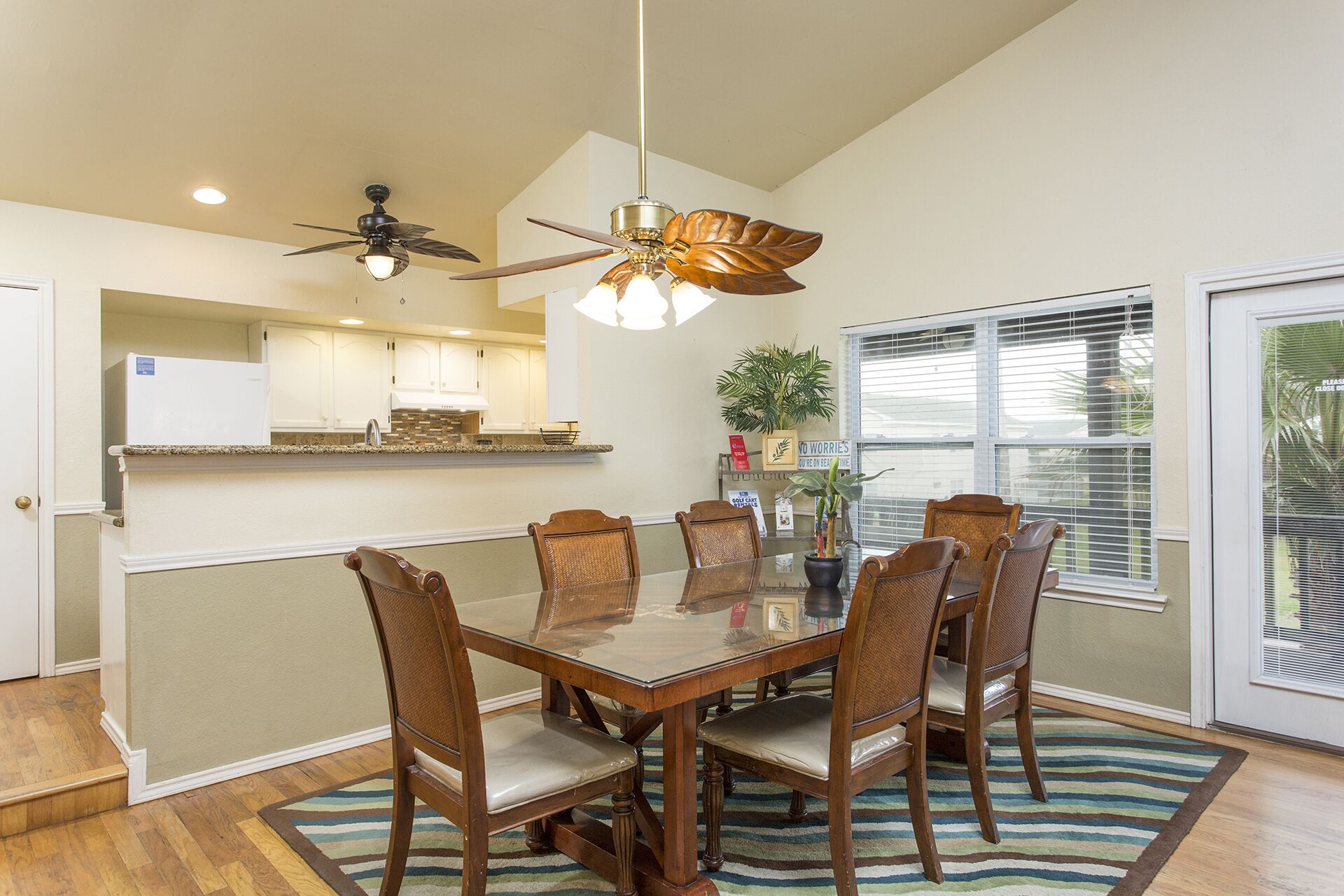 A dining area with a glass table surrounded by wooden chairs, a ceiling fan, and a window with blinds.