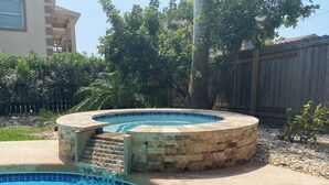 A circular hot tub with a stone exterior and a blue-tiled edge, surrounded by a concrete deck and a wooden fence. - Bahama Beach House • Private Heated Pool & Hot Tub • 3 Min Walk to Beach (South Padre Island)