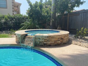 Un jacuzzi rond, avec une structure extérieure en pierre et un bord recouvert de carreaux bleus, entouré d’une terrasse en béton et d’une clôture en bois.
