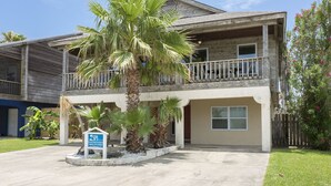 A two-story building with a balcony on the upper floor, surrounded by palm trees and a small sign in front. - Bahama Beach House • Private Heated Pool & Hot Tub • 3 Min Walk to Beach (South Padre Island)