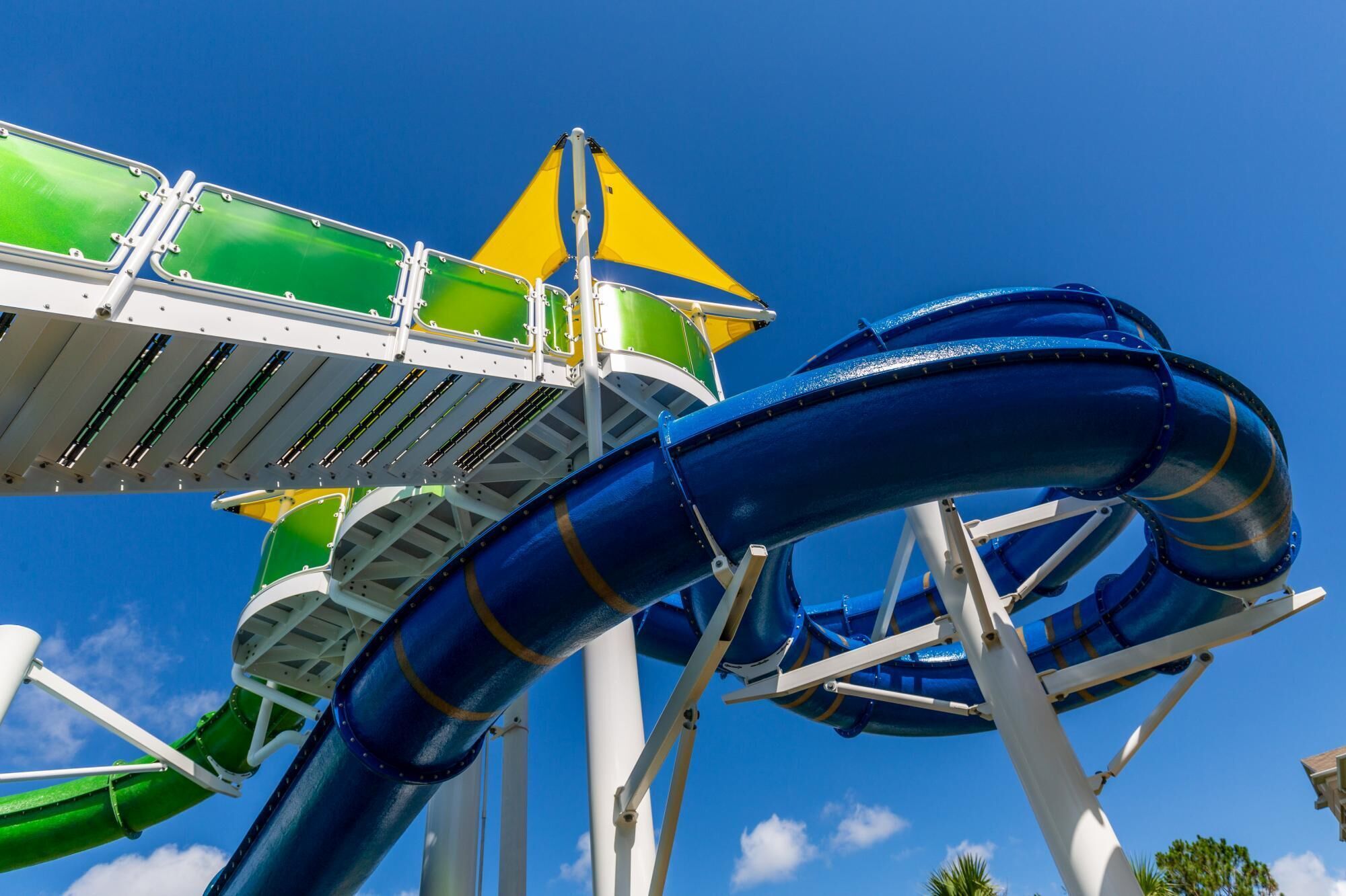 A water slide structure with green and blue tubes, a yellow safety sign, and a clear blue sky.