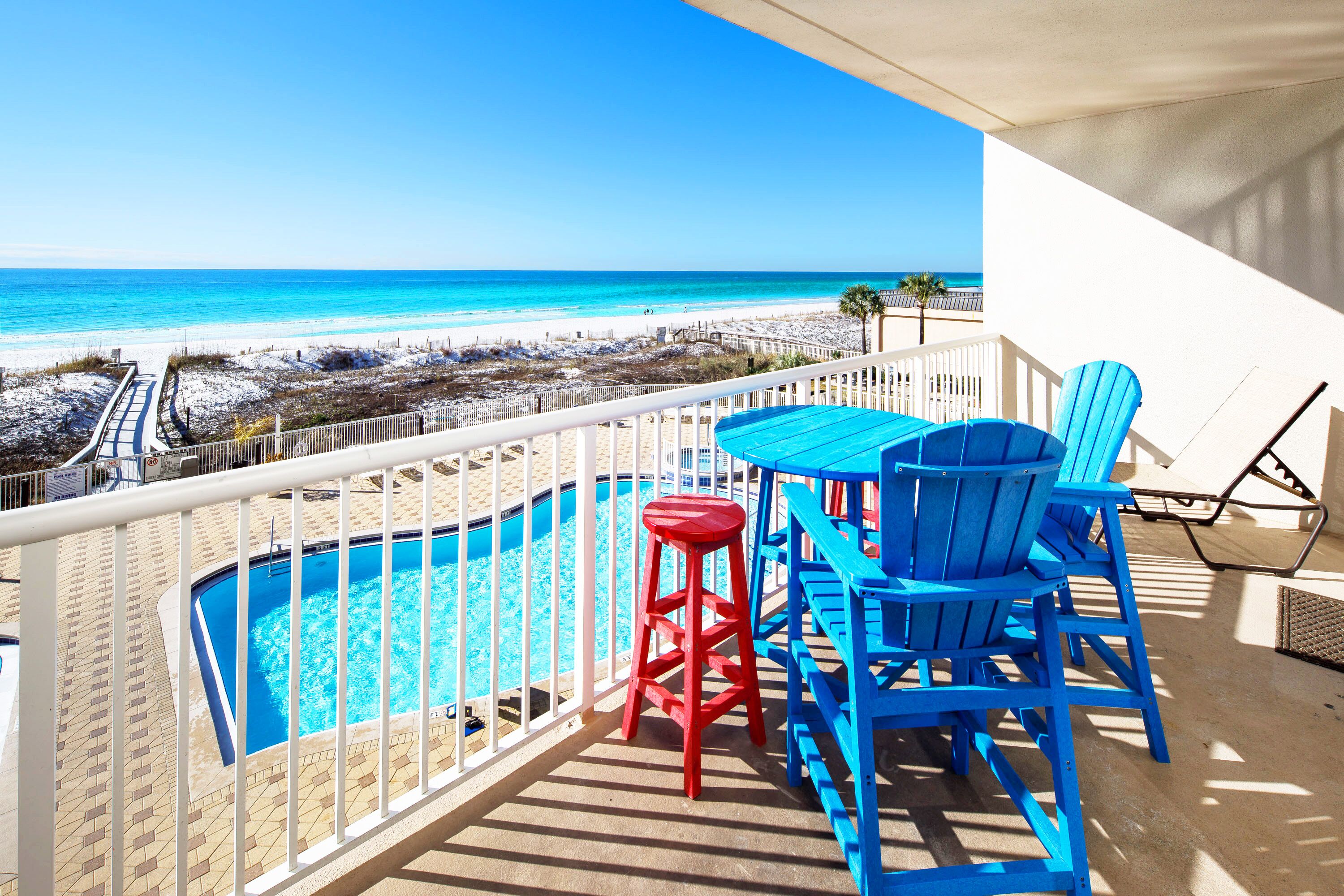 Un balcón con piscina, sillas azules y un taburete rojo con vista a la playa.