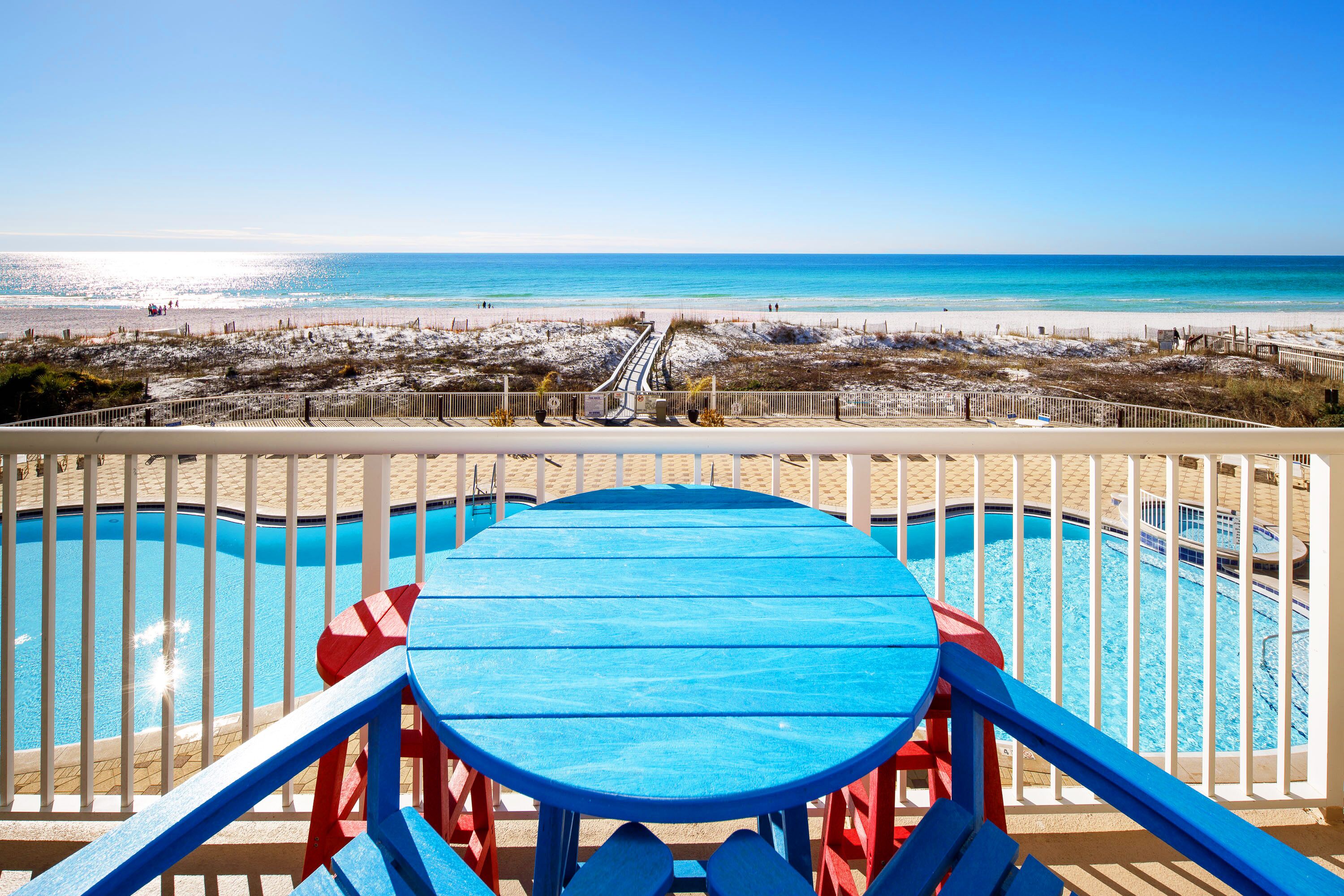 A pool area with a blue table and red chairs, a beach view, and a pathway leading to the beach.
